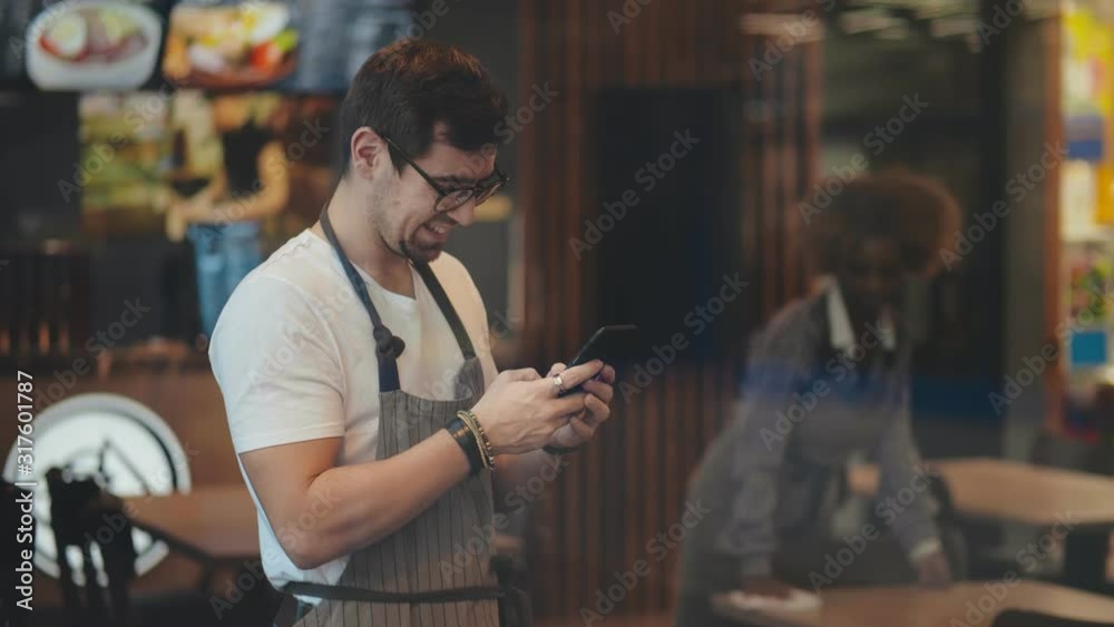 Handsome cafe owner in apron using smartphone ordering supplies online.
