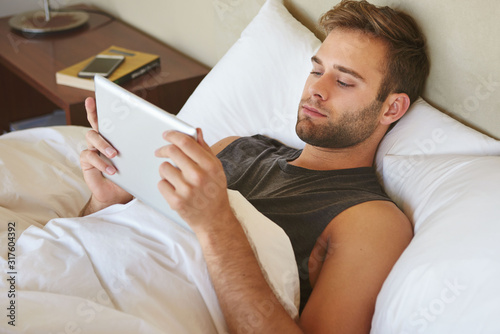 Young man lying in bed reading ebook on digital tablet