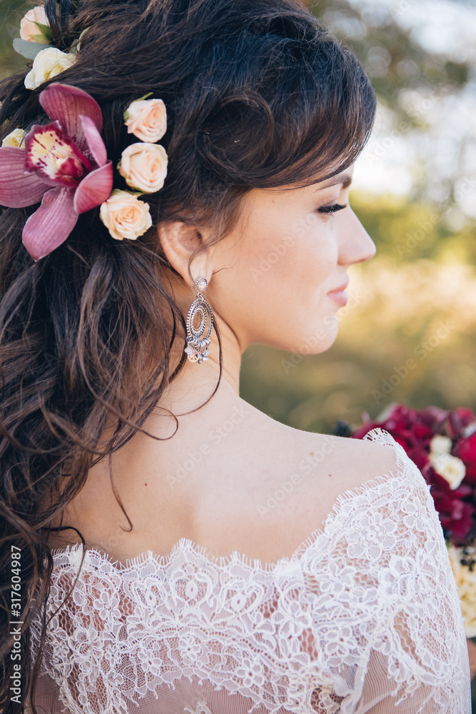 Naklejka premium Closeup portrait of a beautiful girl in a lace dress with a bouquet of red and burgundy flowers in her hands. A young woman walks outdoors, laughs and smiles. Girl with flowers in her hair.