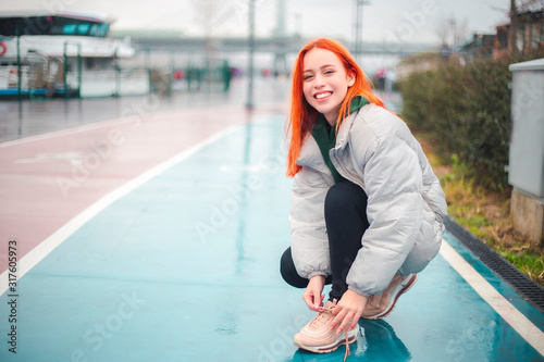 Attractive yound redhead woman lacing her shoes before an exercise, looking to camera and smiling