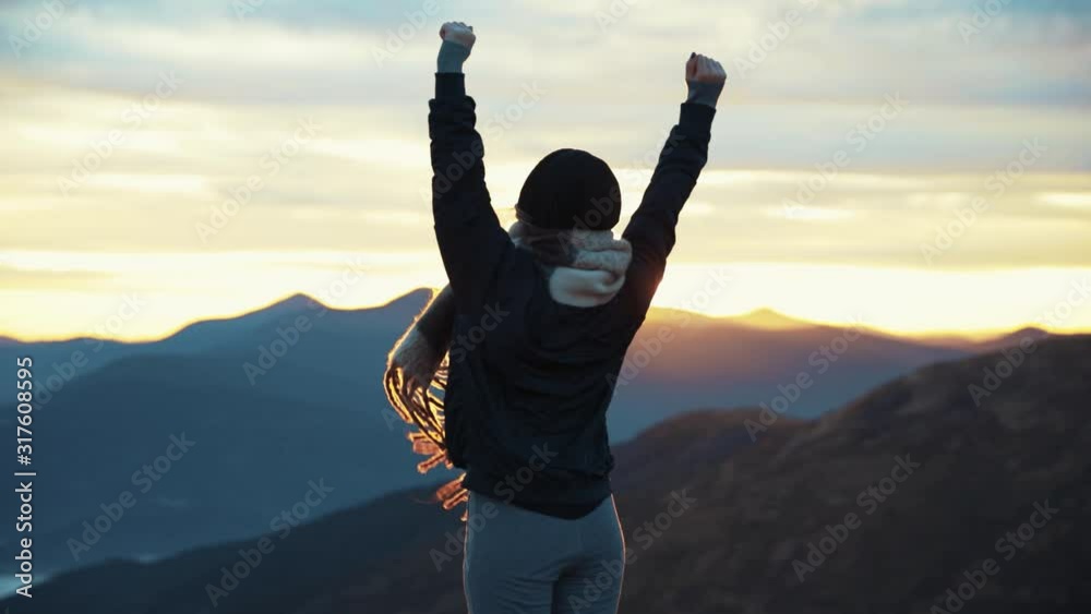 Back view of hipster millennial young woman on top of mountain summit at sunset raises arms into air happy and drunk on life youth and happiness