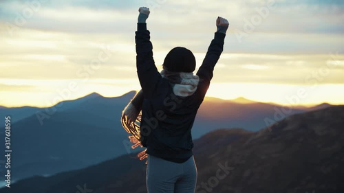 Back view of hipster millennial young woman on top of mountain summit at sunset raises arms into air happy and drunk on life youth and happiness