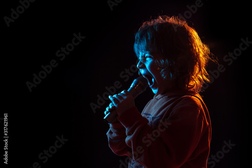 Singing like celebrity, rockstar. Caucasian boy's portrait on dark studio background in neon light. Beautiful curly model. Concept of human emotions, facial expression, sales, ad, music, hobby, dream.