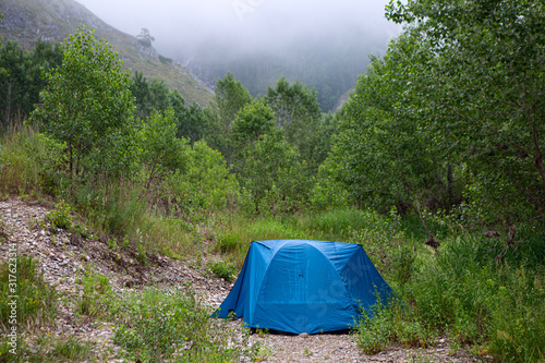 Blue Tourist Tent Or Camp On Background Of Mountain And Green Trees. Early Foggy Morning In Mountainous Place. Concept Of Unity With Nature And Tourism.