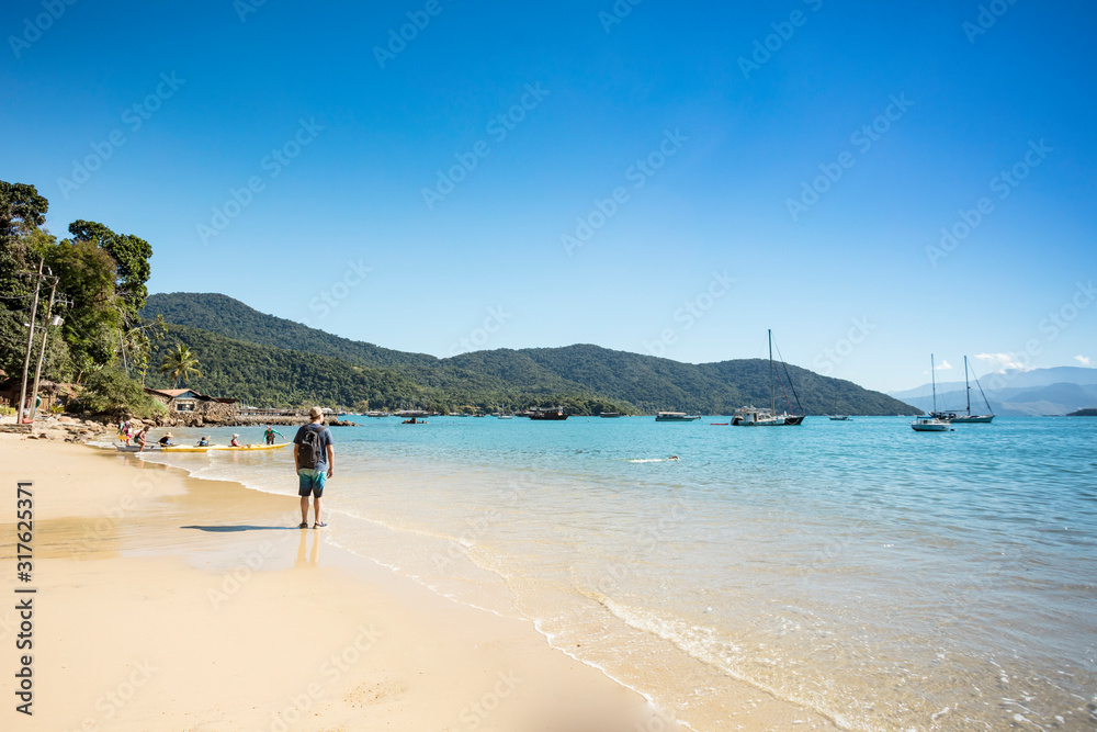 Man walking on the sand of Abraaozinho beach with taxi boat and blue waters in Abraao, on the tropical Ilha Grande, Angra dos Reis, in the south of Rio de Janeiro Brazil