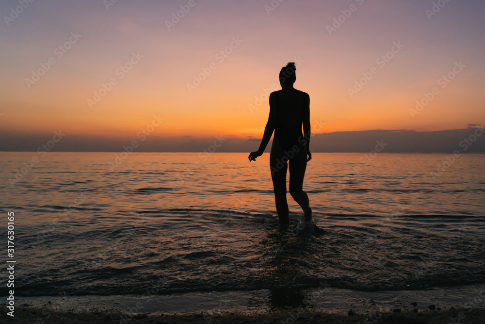 Silhouette of a young slender woman standing in the sea against the background of dawn