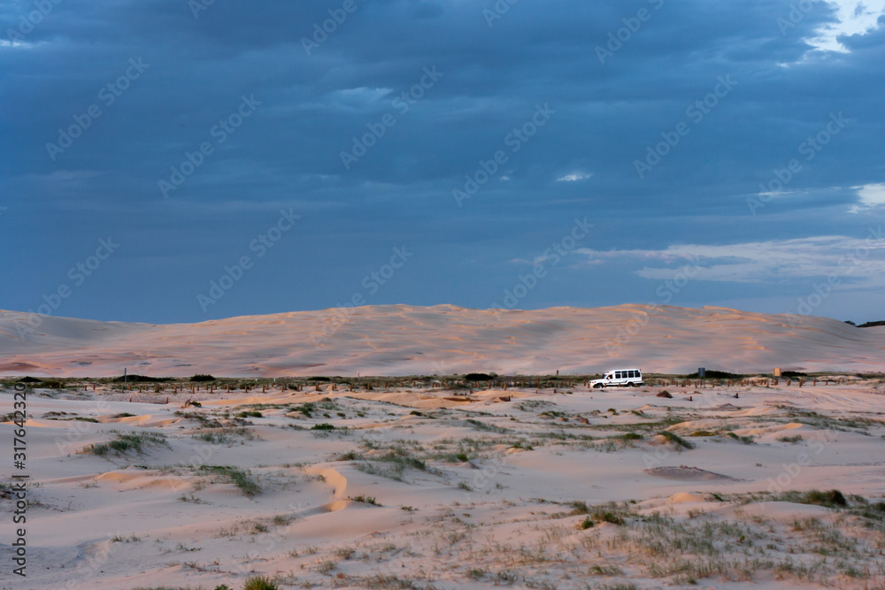 Wide boundless flat desert and a point of a white car driving along horizontal stripe of a road far away. Stockton Sand Dunes near the coast, Worimi Regional Park, Anna Bay, Australia