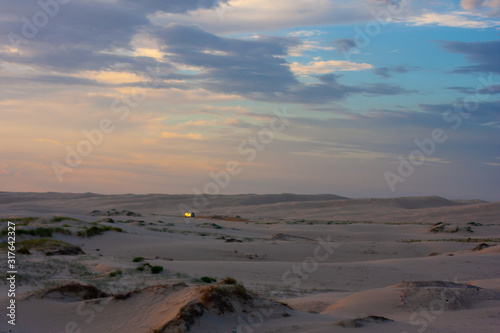 Wide boundless flat desert and a point of a white car driving along it far away. Twilight. Stockton Sand Dunes near the coast, Worimi Regional Park, Anna Bay, Australia