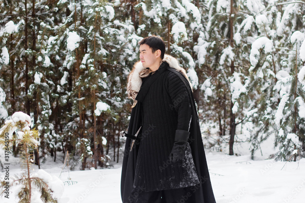 A warrior wearing black quilted clothes in a black cloak with a white fur collar and a long sword in his hands is walking along deep snow against the backdrop of a pine forest.