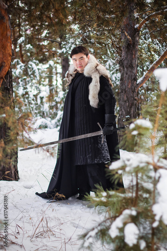 A warrior wearing black quilted clothes in a black cloak with a white fur collar and a long sword in his hands is walking along deep snow against the backdrop of a pine forest.
