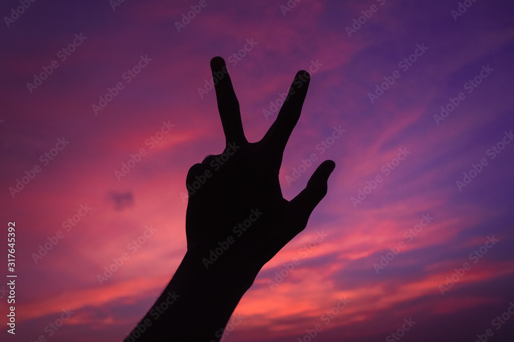 Silhouette of a hand making peace sign in front of the clouds during susnet