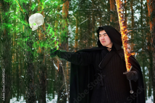 Warrior, magician in black quilted clothes and in a cloak with a hood against the backdrop of a winter forest holds a sword and a staff with a skull in his hands.