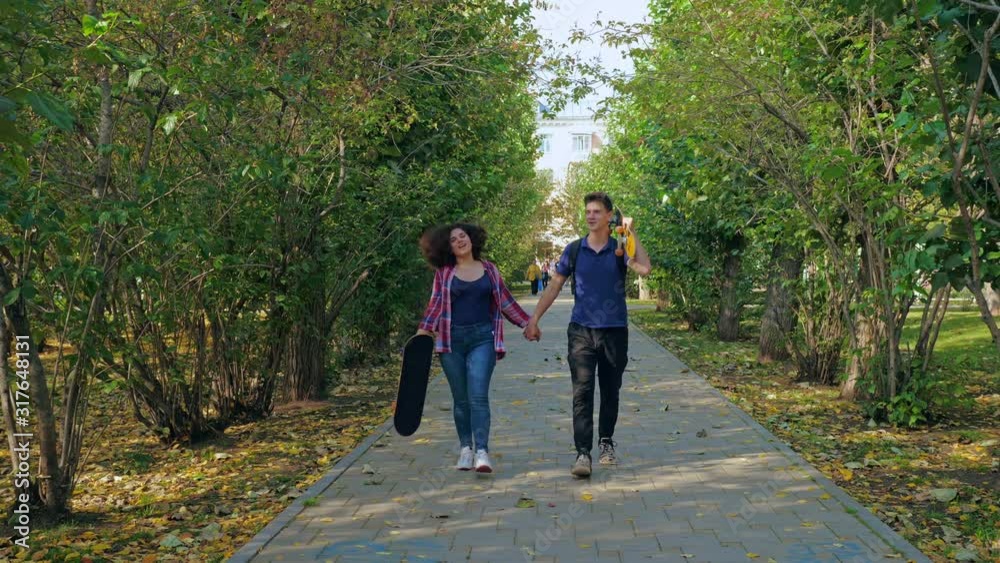 Content young male with skateboard holding hands with optimistic plump girlfriend walking in autumnal park looking away Content young male with skateboard holding hands with optimistic plump girlfriend walking in autumnal park looking away