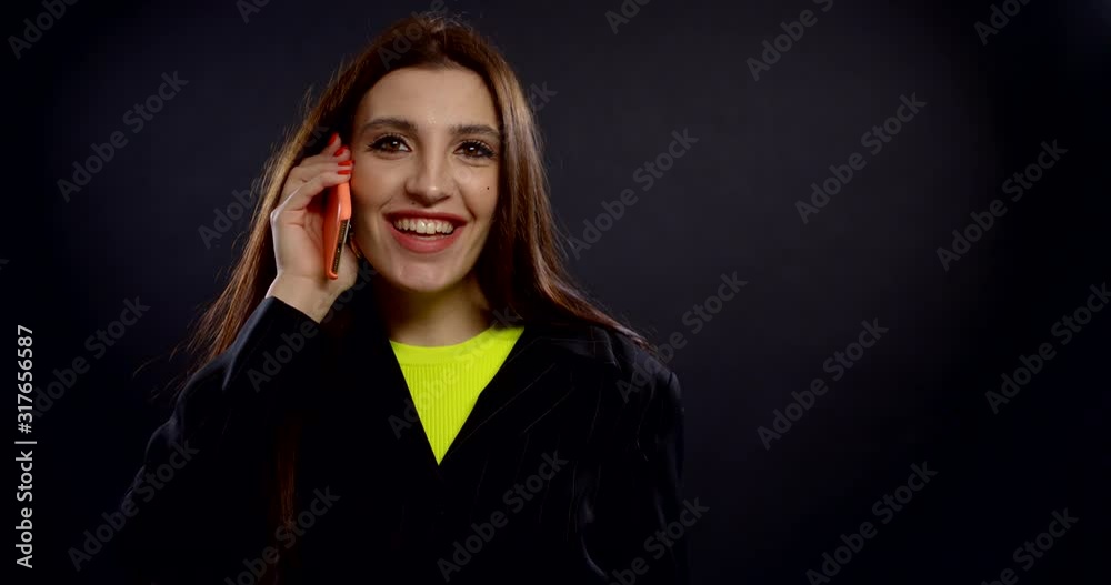 A close-up portrait of a smiling, bright, dark-haired, brown-eyed girl who is in the Studio on a dark background and talking on the phone, wearing a bright yellow t-shirt and a black jacket on top. A close-up portrait of a smiling, bright, dark-haired, brown-eyed girl who is in the Studio on a dark background and talking on the phone, wearing a bright yellow t-shirt and a black jacket on top.