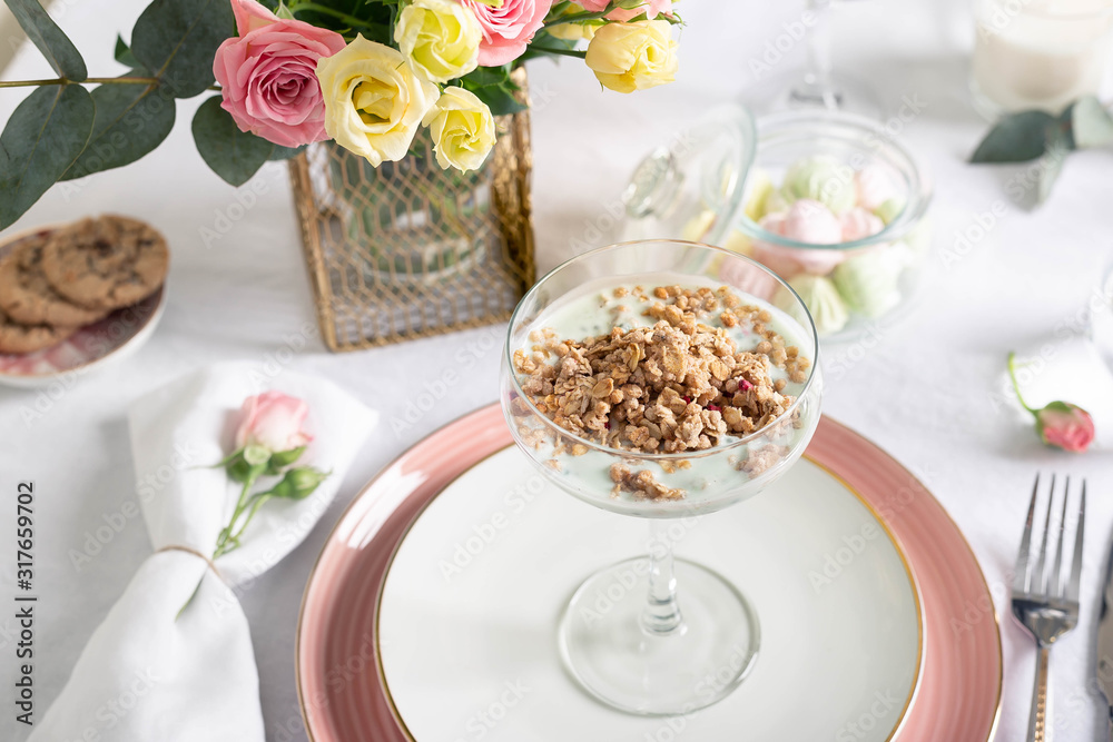 Sunday breakfast at the restaurant, served with granola and Greek yogurt in a glass goblet on porcelain dishes with coffee, cereal cookies and a bouquet of flowers on a white tablecloth.