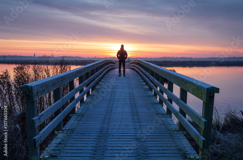 A man on a wooden bridge over a lake enjoying the view during a tranquil sunrise.