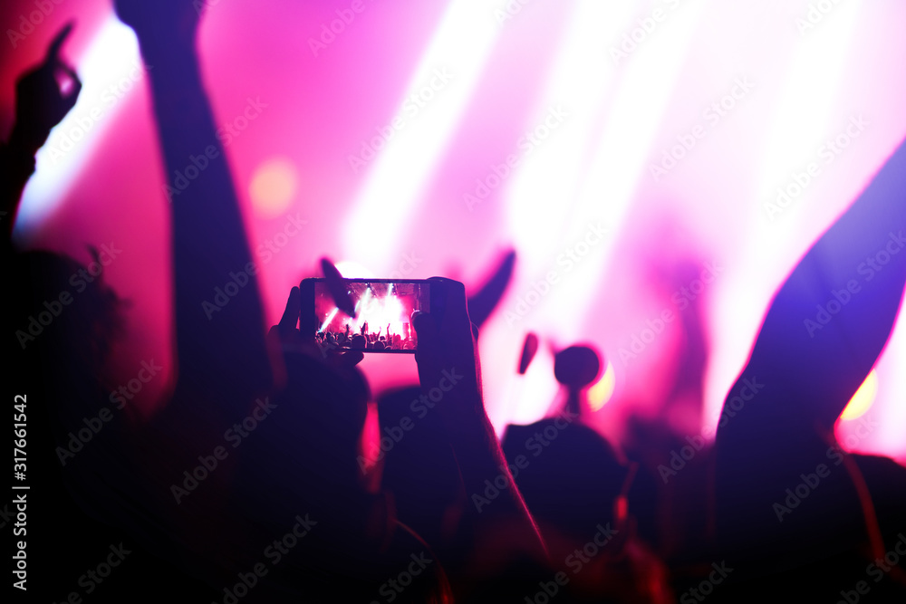 Naklejka premium Hand with a phone records live music festival. People taking photographs with smart phone during a public music concert. Crowd raising their hands, dancing and enjoying great the concert.