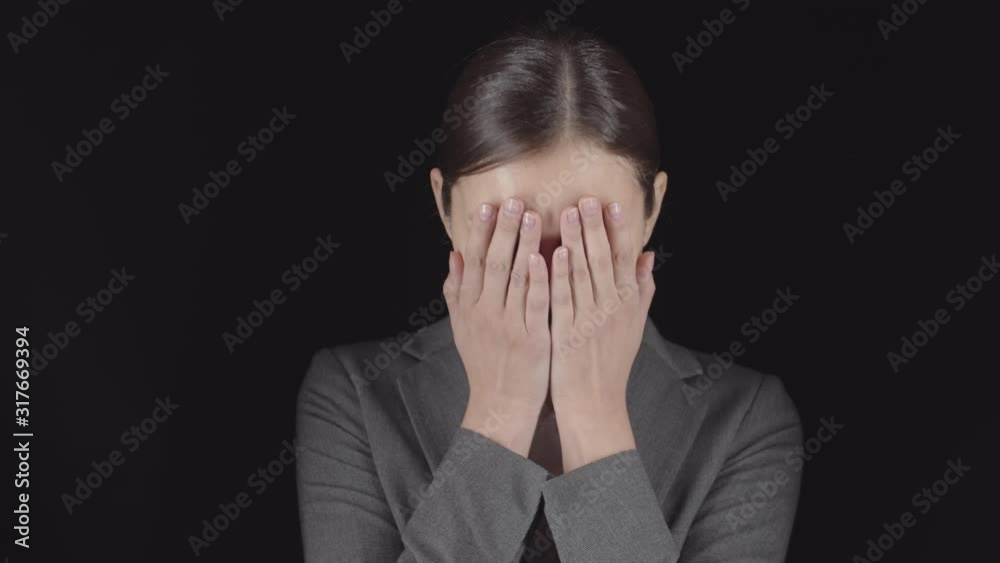 Medium shot of young Caucasian woman wearing casual clothes standing against black wall, looking at camera and then putting palms on her face Medium shot of young Caucasian woman wearing casual clothes standing against black wall, looking at camera and then putting palms on her face