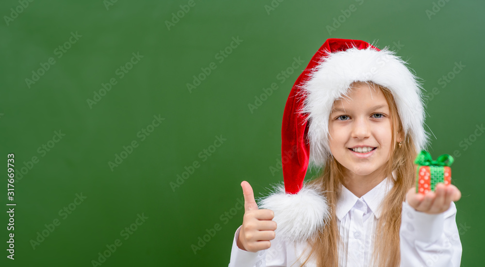 Smiling girl wearing a red christmas hat holds gift box near green chalkboard and shows thumbs up gesture. Empty space for text Smiling girl wearing a red christmas hat holds gift box near green chalkboard and shows thumbs up gesture. Empty space for text