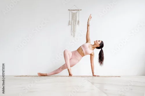 Obraz Young attractive woman practicing yoga in bright yoga class on wooden eco floors tanding in Full Version of Vasisthasana exercise, Side Plank pose, working out, pink sportswear. White wall background