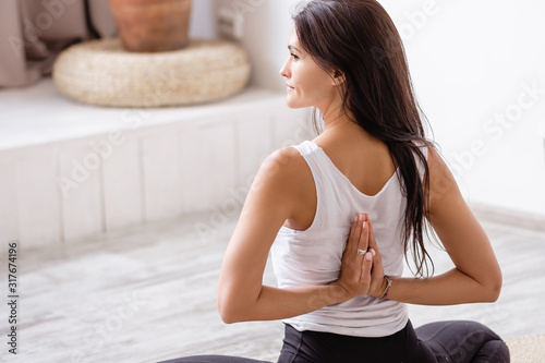 Rear view of an elegant young slim brunette woman doing baddha konasana sitting on a rug on the floor with her eyes closed in cozy quiet environment. Health and Body Improvement Concept. Home fitness photo