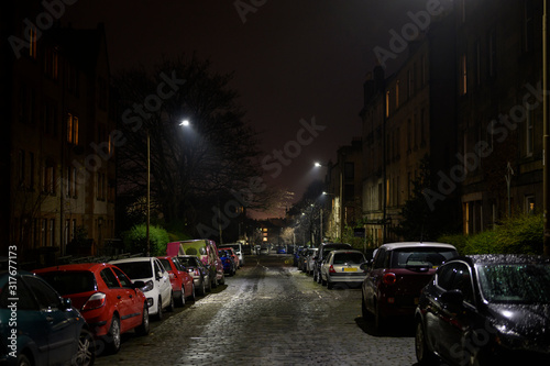 British urban streets at night. Dark alley of Edinburgh city with cars parking on the side of the road, damp from the rain. Downtown apartment buildings and homes neighborhood, street lights on.