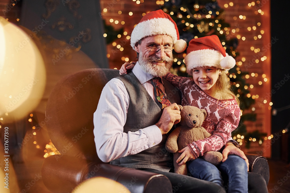 Cheerful fashioned senior man with grey hair and beard sitting with little girl in christmas hats and teddy bear