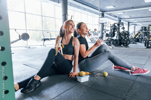 Принти на полотні Two female friends in sportive clothes is in the gym earing fruits and taking a