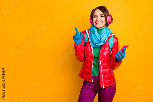 Photo of funny blogger lady hold telephone listen earphones directing finger empty space wear casual red coat blue scarf gloves trousers isolated yellow color background