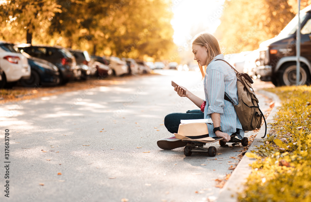 Female tourist sitting on long board with a map in the city and looking at phone. - obrazy, fototapety, plakaty 