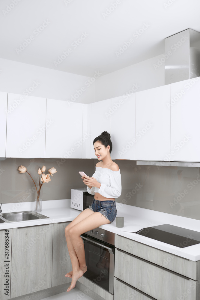 Young beautiful Asian woman using smartphone and smiling while sitting in kitchen Young beautiful Asian woman using smartphone and smiling while sitting in kitchen
