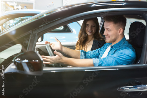 young woman manager in business suit consulting young man client in electric car dealership center, professional cooperation © studioprodakshn