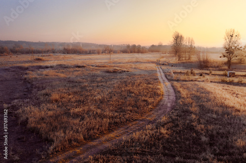 A dirt road extending into the distance among yellowed dry grass in morning meadow. The road through the field in the morning at dawn in the rays of the rising sun. Beautiful landscape.