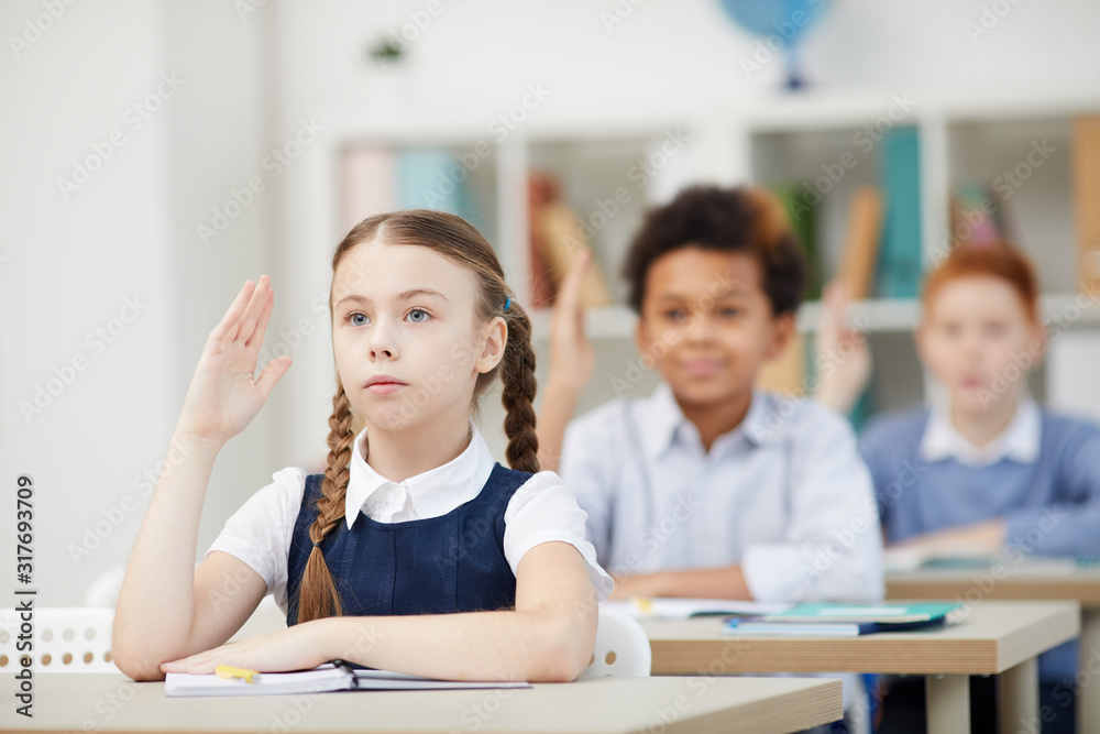 Serious schoolgirl sitting at desk and raising her hand with other classmates in the background during a lesson at school Serious schoolgirl sitting at desk and raising her hand with other classmates in the background during a lesson at school