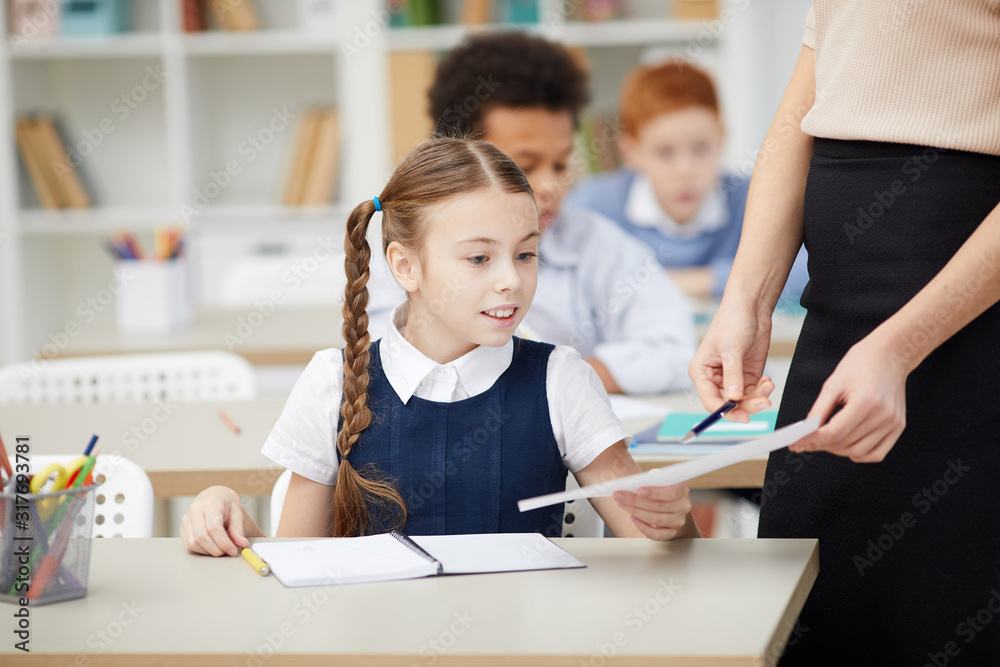 Teacher standing and pointing at paper she showing the work of schoolgirl with good mark during the lesson Teacher standing and pointing at paper she showing the work of schoolgirl with good mark during the lesson