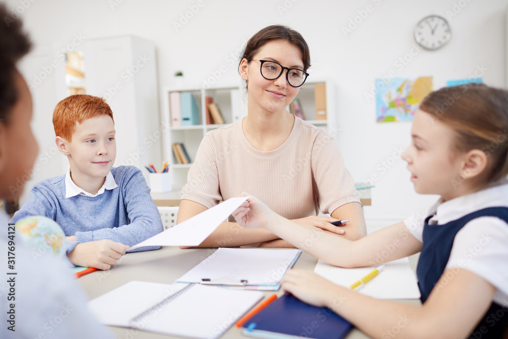 Schoolgirl giving a paper to the boy while they sitting at the table together with their teacher and working in team