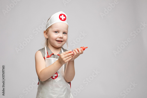 Little cute funny girl playing wearing doctor uniform holding toy syringe looking at camera. Smiling baby female child in glasses posing in nurse garb at white studio background medium long shot