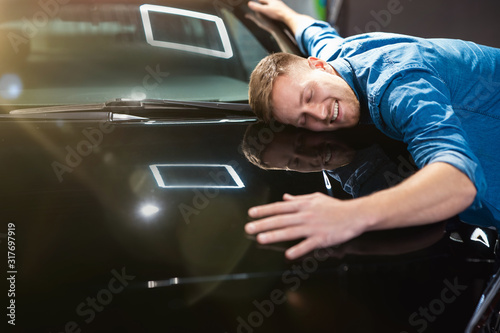 young handsome man feels happy and joyful hugging his brand new vehicle in electric car dealership center, close up