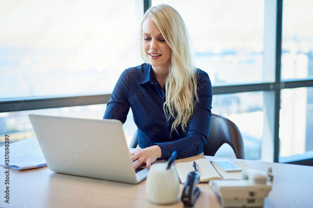 Smiling businesswoman sitting at her office desk using a laptop Smiling businesswoman sitting at her office desk using a laptop