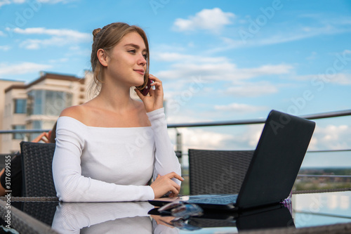 Woman working on laptop at office while talking on phone. Portrait of young smiling business woman calling her best friend, having break, telling something funny, sitting in cafe.