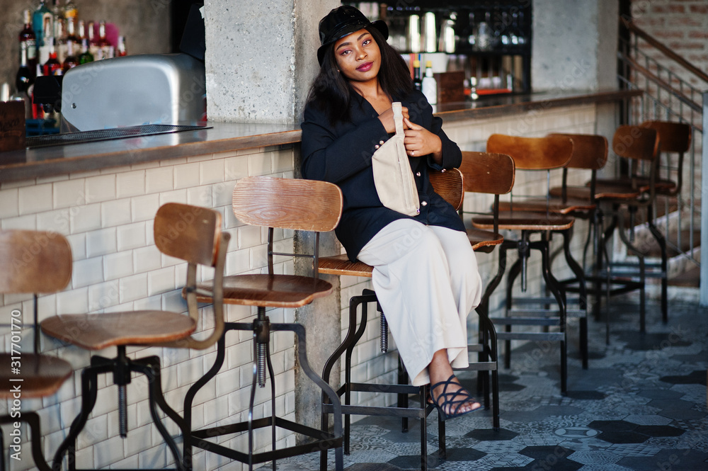 Charming african american woman model in black jacket, hat and waist bag relaxing in cafe during free time.