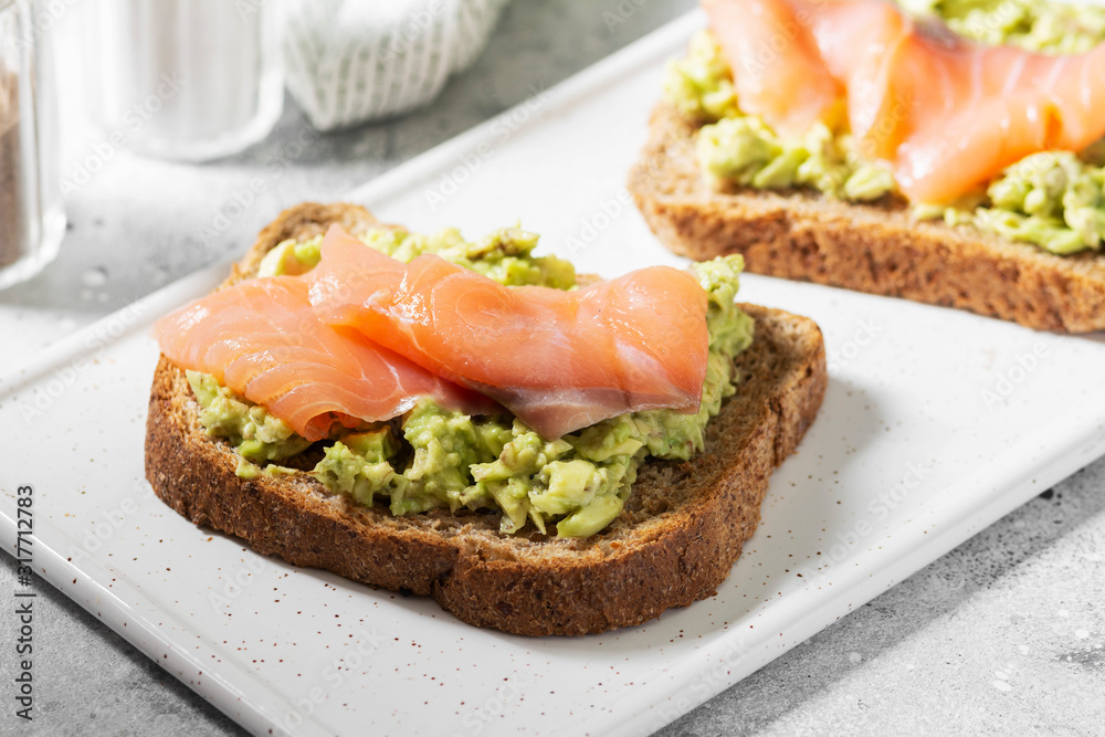 Bruschetta with avocado and salmon on a white ceramic plate on a light gray kitchen table. Toasted bread with avocado and red fish. The concept is helpful and a delicious Breakfast