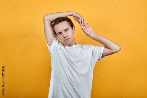 Young man in white t-short on yellow background raises his hand up and squeezes it in lock behind head © Petro