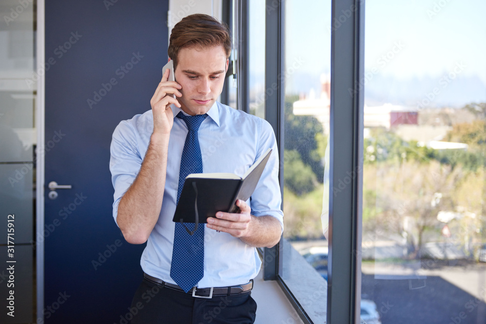 Businessman checking diary while talking on mobile phone in office