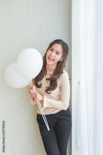Vertical portrait beautiful young happy Asian woman in beige casual wear is smiling and holding white balloons side a white blinds with white wall background.