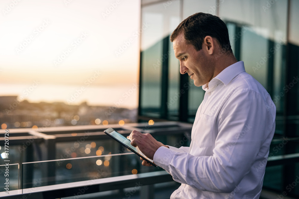 Smiling businessman standing on an office balcony using a tablet - obrazy, fototapety, plakaty 