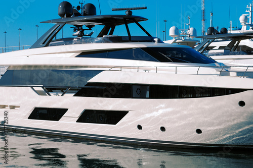 Side view of a part of a luxurious white yacht with dark glasses standing in a bay next to other pleasure yachts.