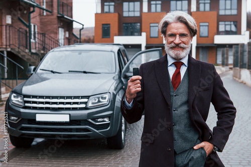 Fashionable senior man with gray hair and beard standing outdoors on the street near his car with keys in hand © standret