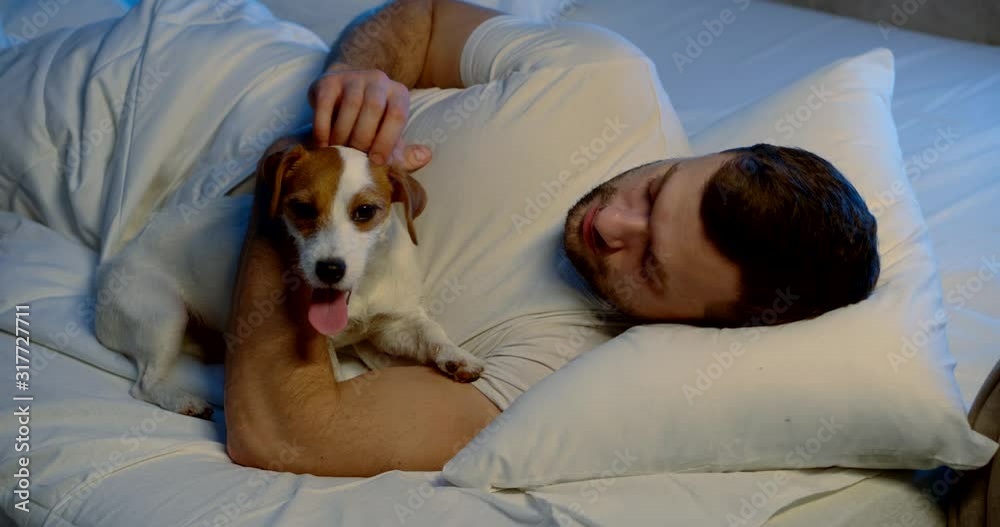 A portrait of a dark-haired man with a middle-aged beard, lying in a white bed with his small dog, playing with it, hugging it, stroking it.