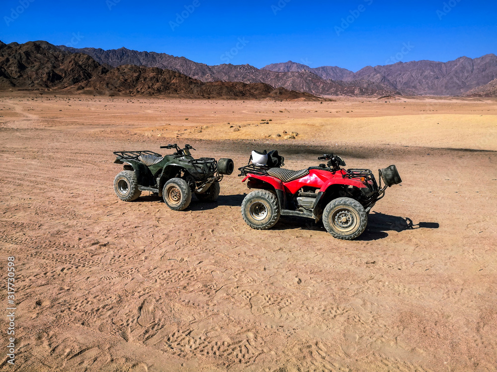 Two ATVs against the backdrop of mountains in the South Sinai Desert near Sharm El Sheikh (Egypt). Red and black quadricycles are standing on the sand, the concept of a tourist excursion on quads Two ATVs against the backdrop of mountains in the South Sinai Desert near Sharm El Sheikh (Egypt). Red and black quadricycles are standing on the sand, the concept of a tourist excursion on quads